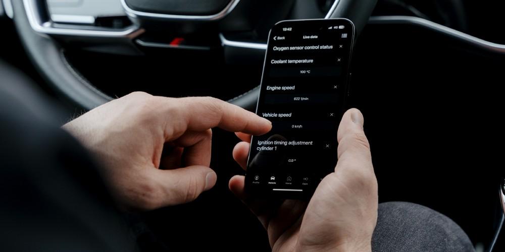 A person is holding a smartphone displaying an OBDeleven OBD2 scanner app with live sensor data from a vehicle. Visible readings include oxygen sensor data, coolant temperature, engine speed, vehicle speed sensor data, and ignition timing adjustment data.