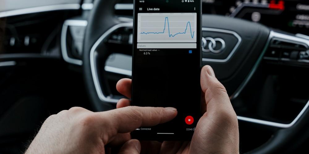  A person holding a smartphone inside a car, with an OBDeleven OBD2 scanner app open displaying live sensor data, including a chart of the calculated engine load and its numerical value from the engine control module.