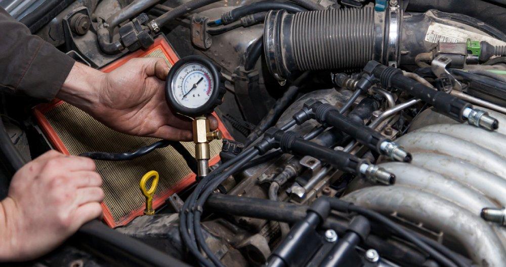 A person is performing an engine compression test, holding a pressure gauge connected to a car engine cylinder.