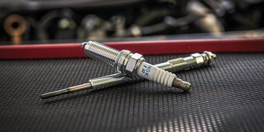 Spark plug and glow plug positioned against a textured black background with a red border, suggestive of a mechanic's work mat.