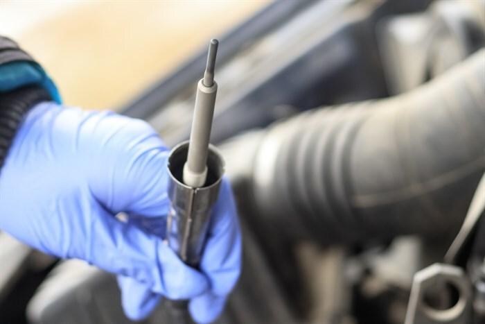 A hand in a blue glove holds a metal glow plug socket wrench with a glow plug inserted, ready for installation