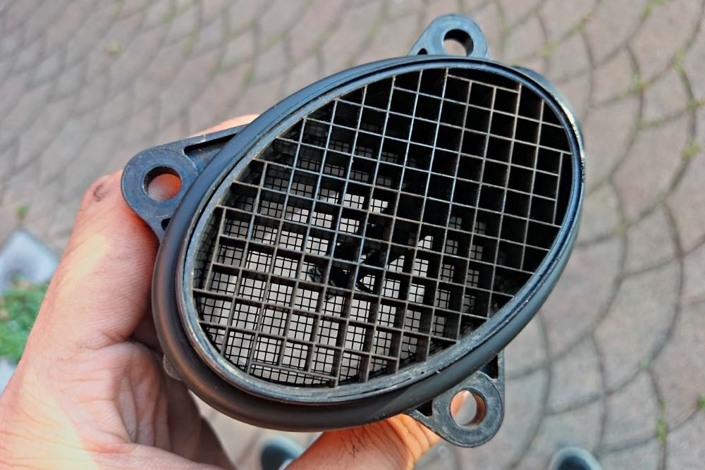 A close-up view of a mass airflow (MAF) sensor, showing its plastic housing and internal grid structure, held in hand.