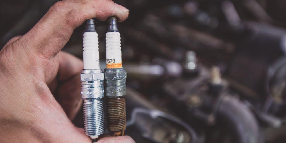  A close-up of a person's hand holding two spark plugs in front of an out-of-focus car engine, with one spark plug appearing new and the other showing signs of wear.