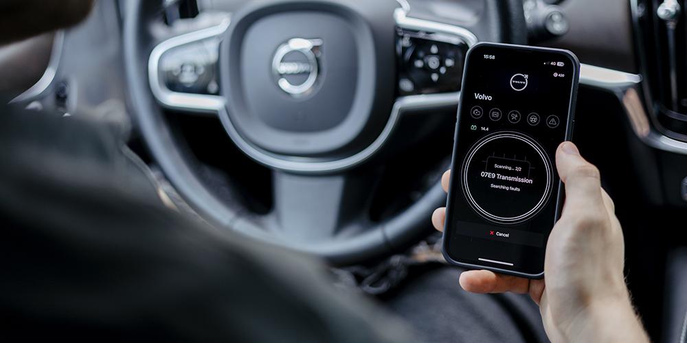 A person in the driver's seat of a Volvo car is holding a smartphone with an OBDeleven diagnostic application and scanning Transmission control unit