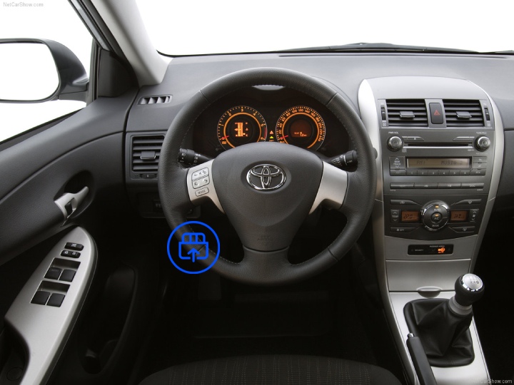 The image shows the interior of a Toyota Corolla, highlighting the OBD-II port location beneath the dashboard near the driver’s footwell.