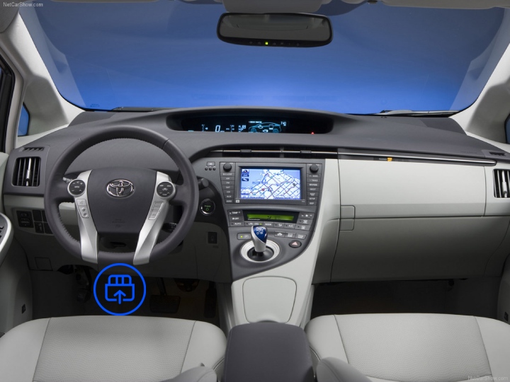 The image shows the interior of a Toyota Prius, highlighting the OBD-II port location beneath the dashboard near the driver’s footwell.