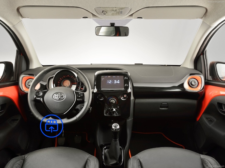 The image shows the interior of a Toyota Aygo, highlighting the OBD-II port location beneath the dashboard near the driver’s footwell.