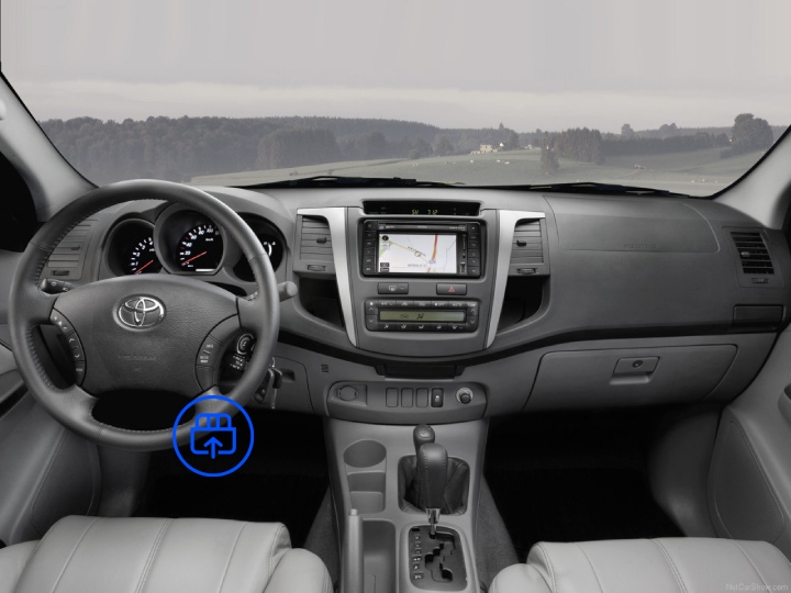 The image shows the interior of a Toyota Hilux, highlighting the OBD-II port location beneath the dashboard near the driver’s footwell.