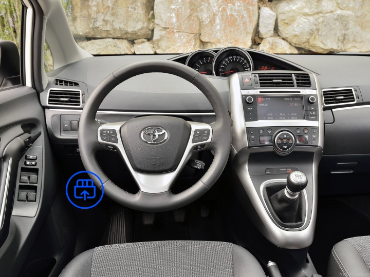 The image shows the interior of a Toyota Verso, highlighting the OBD-II port location beneath the dashboard near the driver’s footwell.